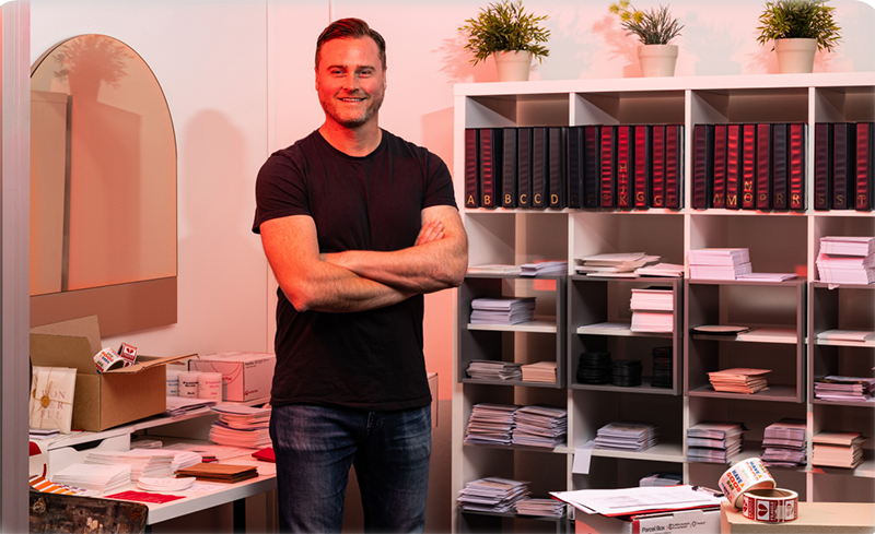 James Boston, Co-Founder of Paperlust, stands in his store, arms crossed and smiling. Stationery and Australia Post boxes are around him.