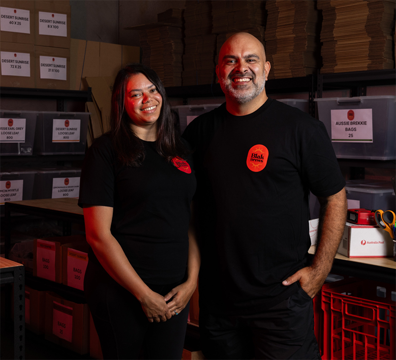 Cerisa Benjamin and Troy Benjamin, Co-Founders of Blak Brews, stand in their warehouse, smiling. Teas and Australia Post boxes are around them.