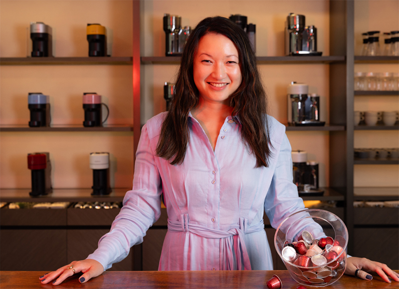 YC Eu, Head of eBusiness at Nespresso, stands in Nespresso's coffee corner, smiling. Nespresso coffee machines and pods are around her.